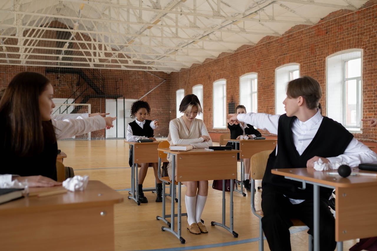 Teenagers in school uniforms pointing at classmate indoors, highlighting bullying.